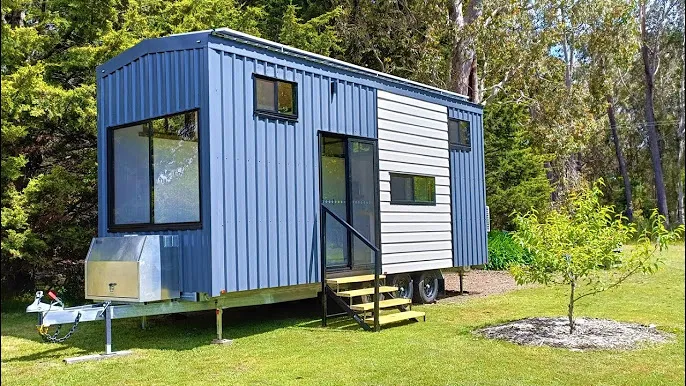 White tiny home with triple axles parked in a wooded area, designed by leading tiny house companies.