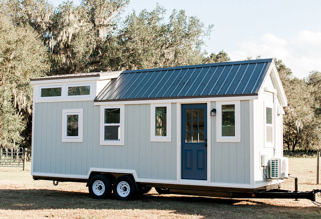 Blue-gray tiny house on wheels parked in a natural setting with trees in the background.